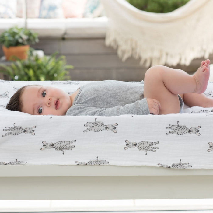 A baby in a gray onesie rests on a zebra print changing pad, surrounded by lush greenery and a hammock. The scene is serene, complemented by aden + anais essentials cotton muslin swaddle blankets from their 4-pack, enhancing the gentle and natural ambiance.