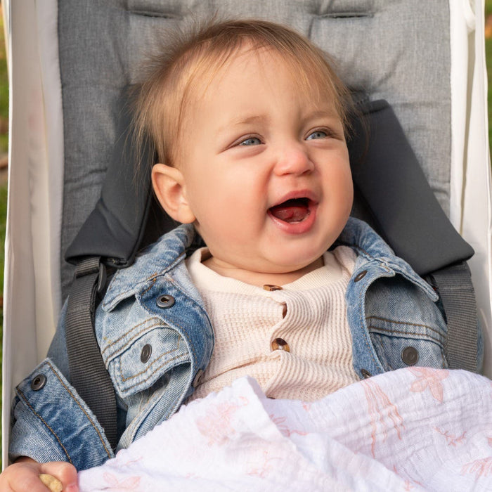 A joyful baby with light brown hair sits in a stroller, wearing a light sweater and denim jacket. The babys laughter fills the air, eyes sparkling above an aden + anais essentials cotton muslin swaddle blanket draped over their lap.
