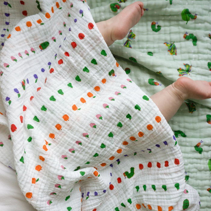A baby’s legs peek out from an aden + anais essentials cotton muslin swaddle blanket, featuring colorful dots and hearts. The baby lies on a frog-print blanket.