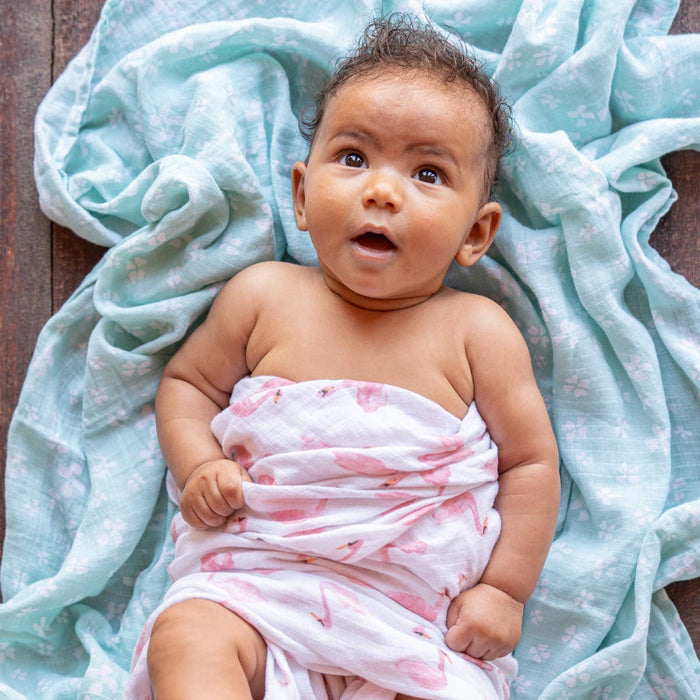 A baby with curly hair lies on a soft, patterned blue blanket, comfortably wrapped in an aden + anais essentials cotton muslin swaddle blanket from the 4 pack with pink designs, gazing upward with an open mouth expression while resting on the wooden floor.