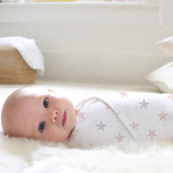 A baby is cozily wrapped in the aden + anais essentials cotton muslin swaddle blanket, lying on a soft cream-colored rug. The softly lit room features a basket and a window in the background.