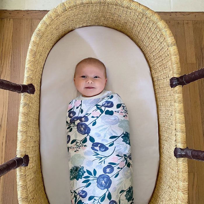A baby lies calmly in a woven basket bassinet on a wooden floor, wrapped in an aden + anais essentials cotton muslin swaddle blanket featuring a floral pattern.