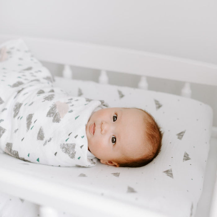 A baby is peacefully swaddled in a breathable essentials cotton muslin blanket by aden + anais, featuring gray and green triangle patterns. The infant lies on a matching changing pad with a calm expression against a soft, neutral background.