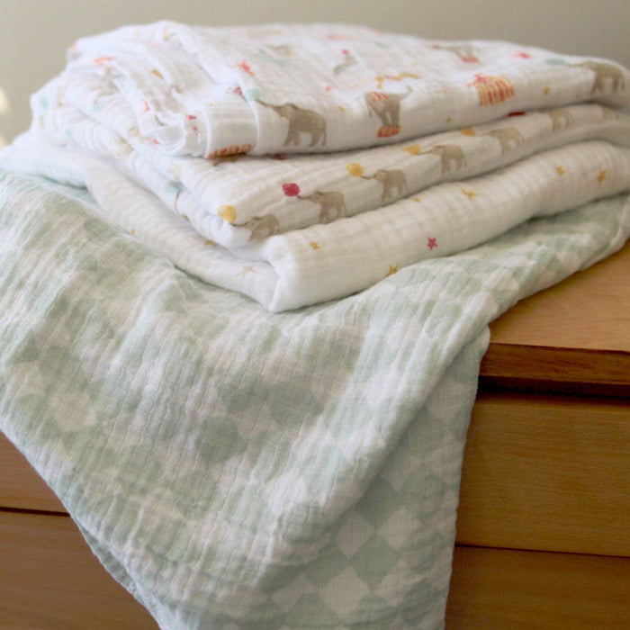 A stack of aden + anais essentials cotton muslin swaddles 4 pack, featuring soft blankets with playful animal designs and a light green geometric pattern, rests on a wooden surface, exuding warmth and comfort.