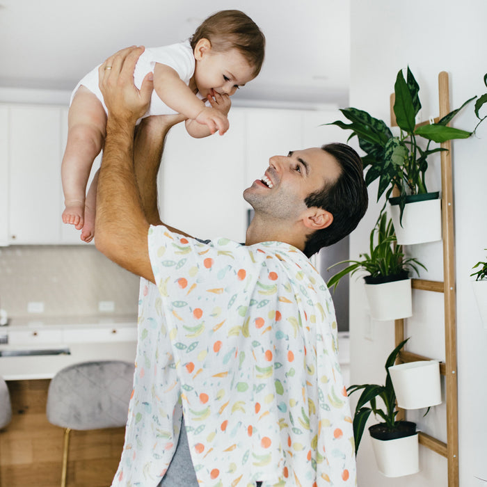In a bright kitchen, a person joyfully lifts a smiling baby wrapped in an aden + anais essentials cotton muslin swaddle blanket. The person wears a patterned cape, while plants adorn a ladder shelf in the background.