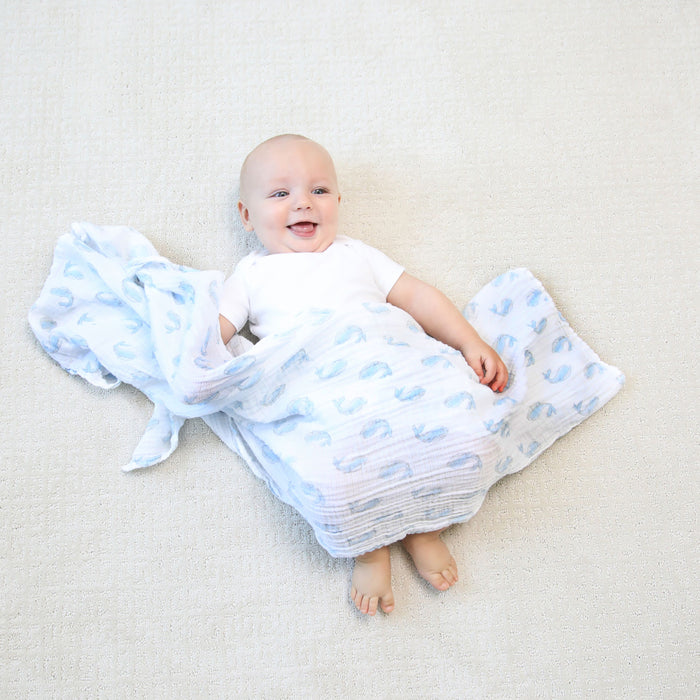 A smiling baby lies on a light-colored surface, wrapped snugly in an aden + anais essentials cotton muslin swaddle blanket. The breathable cotton swaddle features whale or abstract designs, while the baby wears a plain white onesie, showcasing its versatility as a must-have baby essential.
