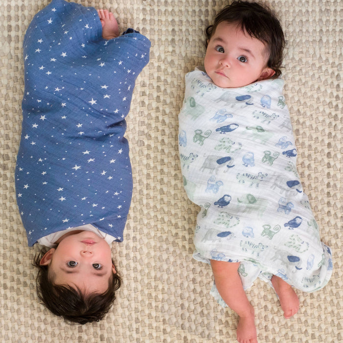 Two babies lie on a textured beige surface, comfortably wrapped in aden + anais essentials cotton muslin swaddle blankets. The left baby is in a breathable blue blanket with stars, while the right is wrapped in white with animal prints. Both have dark hair and gaze upward.