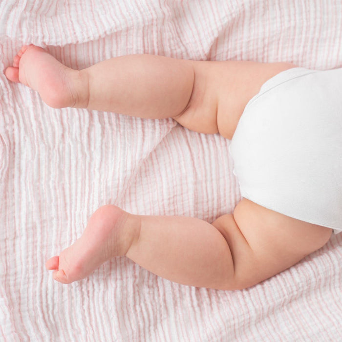 A baby in a white diaper lies on a textured, light pink blanket, with chubby legs and slightly curled feet. The essentials cotton muslin swaddle blanket 4 pack by aden + anais provides breathable cotton fabric and gentle colors, creating a warm and cozy atmosphere.