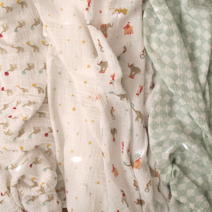 Close-up of three soft, crinkled cotton muslin fabrics from aden + anais. One features circus animals and stars, another circus tents and elephants, with the third a green and white checkered pattern. These versatile Essentials 4 packs make perfect muslin swaddles.