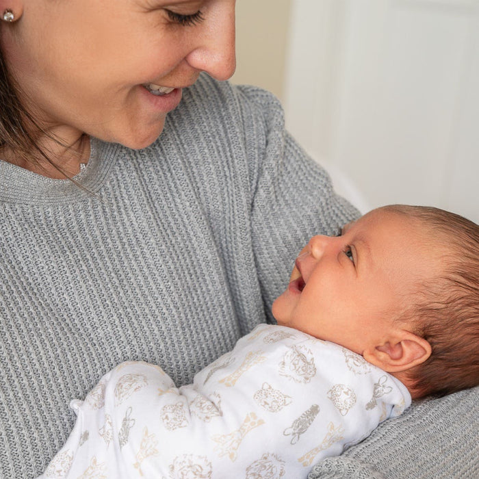 A woman in a gray sweater smiles at her smiling baby swaddled in an aden + anais essentials easy swaddle wrap. They face each other warmly, while the adjustable fasteners ensure startle reflex prevention for the little one.