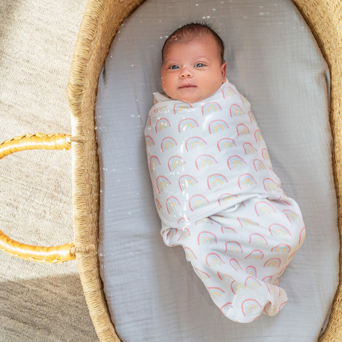 A baby rests in a woven basket, wrapped snugly in an aden + anais essentials easy swaddle wrap, designed to prevent startle reflex. The white wrap with colorful rainbow patterns hugs the little one securely, as wide eyes and a slight smile appear against the soft gray sheet lining the cozy nest.