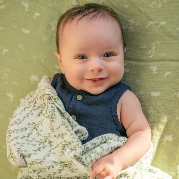 A cute baby with light hair and blue eyes smiles while lying on an aden + anais essentials silky soft swaddle blanket in light green with a plant pattern. The sleeveless navy outfit enhances charm, as the cozy, leafy-patterned blanket is perfect for sensitive skin.
