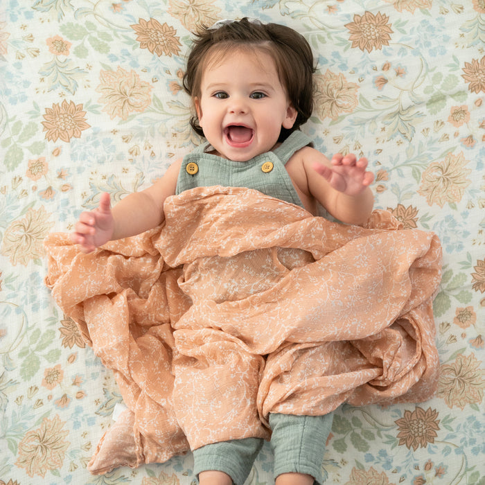 A joyful baby lies on a floral-patterned blanket in a light green outfit, wrapped in aden + anais essentials silky soft swaddle blankets, peach-colored with white patterns. The background showcases soft floral designs.