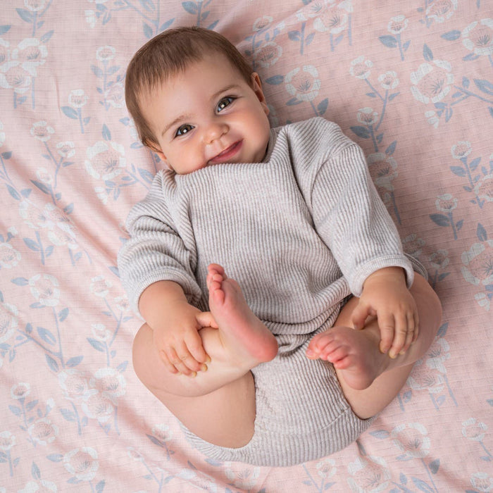 A joyful baby with sensitive skin lies on an aden + anais essentials silky soft swaddle blanket, featuring a pink floral design. They wear a cozy gray outfit and smile while holding their feet, amidst a charming backdrop of light blue and white flowers.