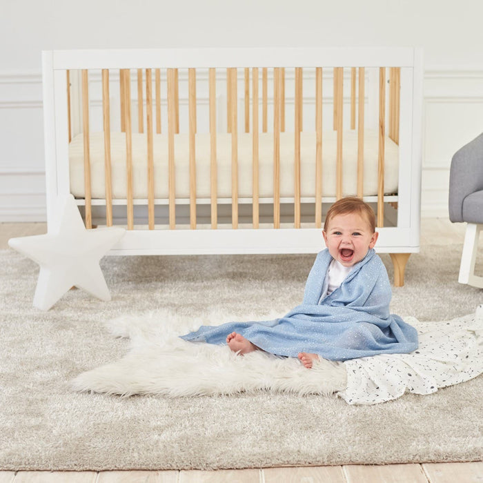 A baby wrapped in an aden + anais essentials silky soft swaddle blanket sits smiling on a fluffy white rug. A white crib with wooden bars, a star-shaped decor, and a gray chair are in the background on the light carpeted floor, ideal for babys sensitive skin.