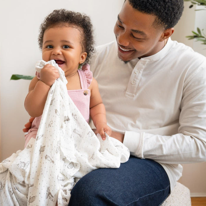 A smiling baby in a pink outfit chews on an aden + anais essentials silky soft swaddle blanket while sitting on an adults lap. The adult, in a white shirt and jeans, smiles warmly at the baby. Green plants create a serene setting ideal for embracing sensitivity.