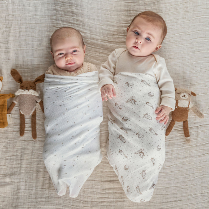 Two babies lay side by side on a soft, light surface, swaddled in aden + anais essentials silky soft swaddle blankets, ideal for their sensitive skin. They look content amidst plush animal toys, including a rabbit on the left and a bear on the right.