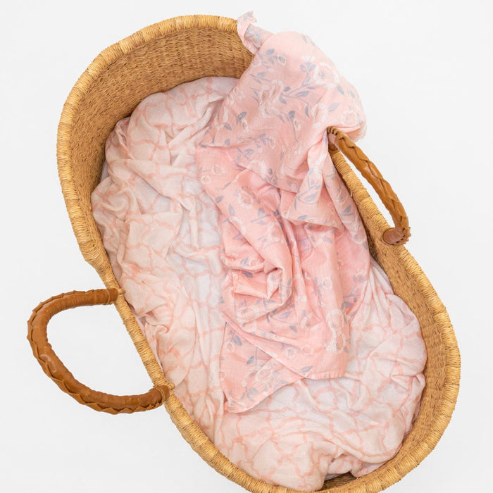A woven basket with leather handles, lined with pink and white fabric, holds an aden + anais essentials silky soft swaddle blanket 2 pack. Perfect for babys sensitive skin, this basket rests elegantly against a white background.