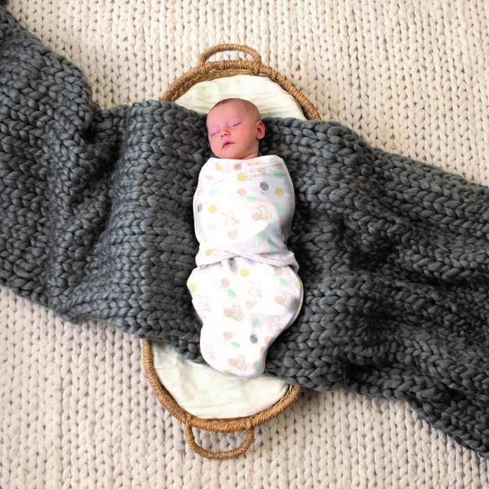 A sleeping baby wrapped in a Disney™ essentials swaddle wrap quick change by aden + anais with pastel patterns lies in a small woven basket on a large gray knitted blanket over a textured beige surface.