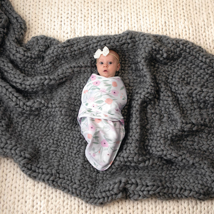 A baby lies on a gray knit blanket in an aden + anais essentials wrap swaddle from the quick change 5 pack, featuring adjustable fasteners for easy changes. The babys surprised expression is accentuated by a white headband with a bow against the cream-colored knitted background.