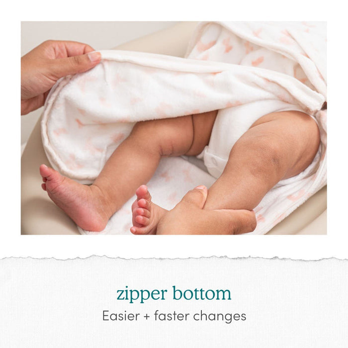 Close-up of a baby on a changing pad, partially wrapped in an aden + anais Disney™ essentials swaddle wrap with a pink pattern. An adult hand unzips the bottom, revealing the babys legs. Text reads: zipper bottom: Easier + faster changes with adjustable fasteners.
