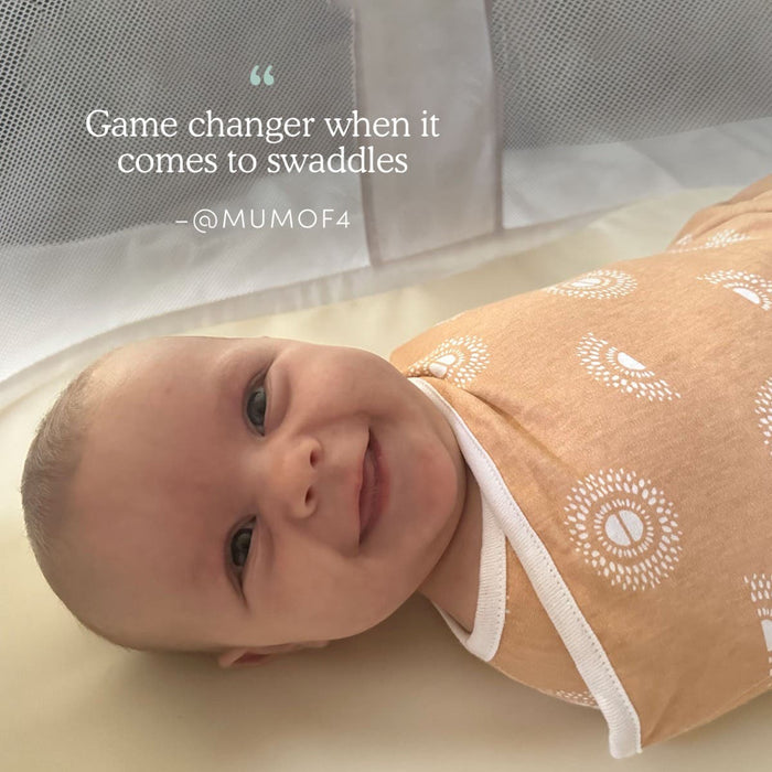 A smiling baby wrapped in an aden + anais Disney™ Winnie the Pooh essentials swaddle wrap quick change lies on a white sheet. Text above reads, “Game changer when it comes to swaddles — @MUMOF4”.