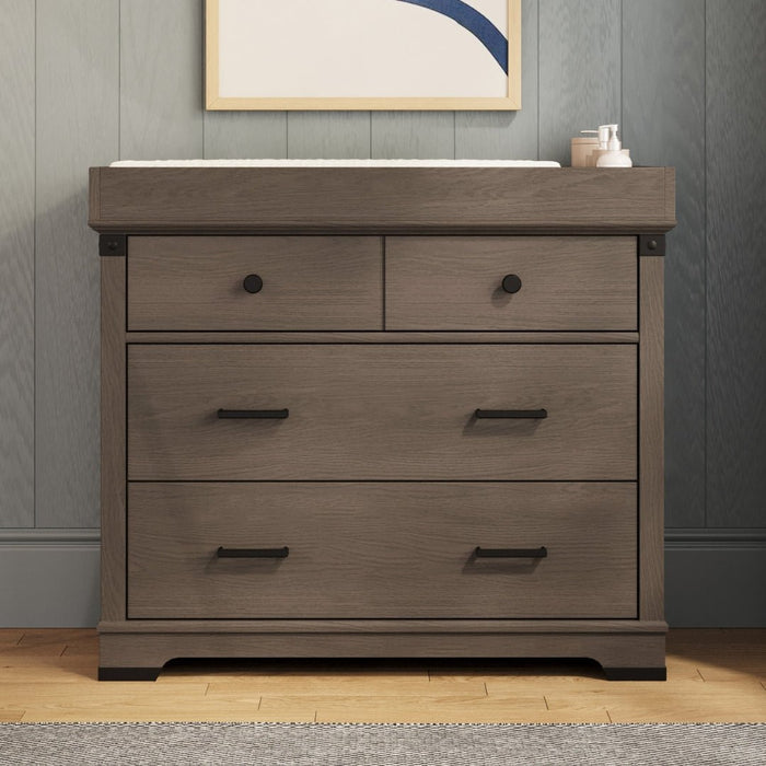 A dark wooden dresser with black handles sits against a gray paneled wall topped with a framed abstract print. The right side holds items—ideal for adding the Child Craft F01792 Universal Changing Table Topper in Dapper Gray.