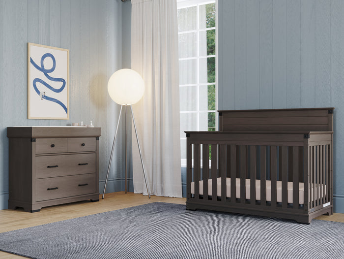 A modern nursery with blue walls, dark wood crib, matching 3-drawer dresser featuring the Child Craft Universal Changing Table Topper in Dapper Gray (F01792), a floor lamp, white-curtained window, gray rug, and abstract wall art.