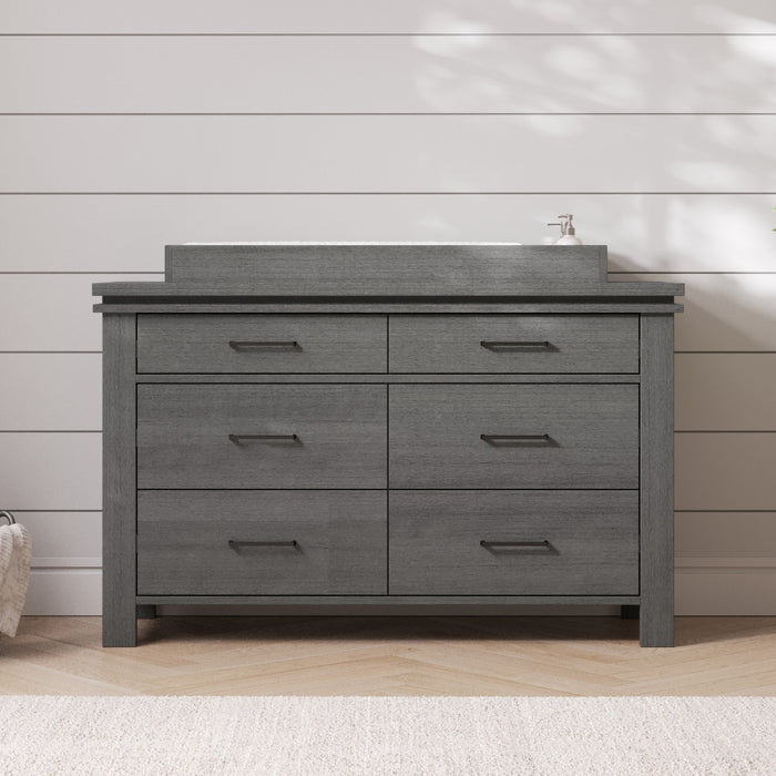 A modern bathroom vanity in midnight gray with six sleek drawers, inspired by the Child Craft F01792 Universal Changing Table Topper, is set against a white shiplap wall, topped with a rectangular sink and bathed in soft sunlight.