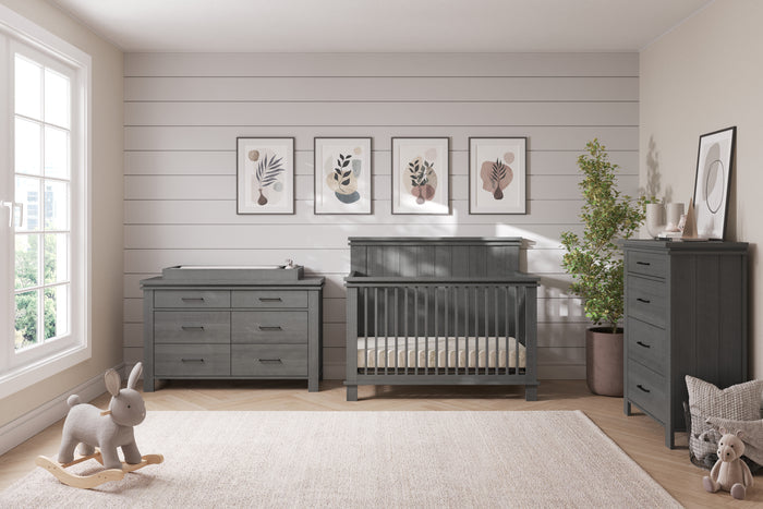 A modern nursery with gray furniture, including a crib, Child Craft dresser, and the F01792 Universal Changing Table Topper in Midnight Gray. Décor features a rocking bunny, plant, framed art on white shiplap walls, and sunlight on a beige rug.