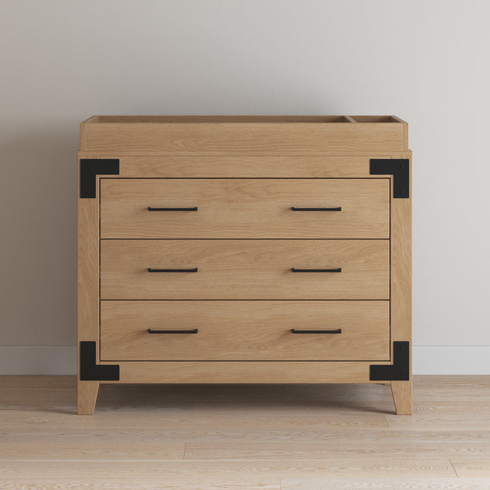 A wooden dresser with three drawers, black metal handles, and corner brackets stands on a light wood floor. Add the Child Craft F01792 Universal Changing Table Topper in Nutmeg to easily convert it into a nursery essential.
