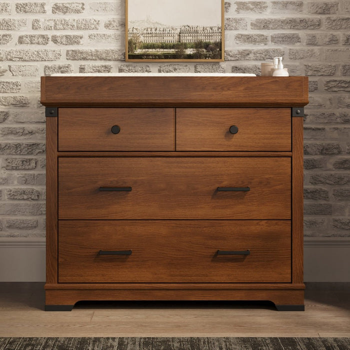 A Child Craft F01792 Universal Dressing Kit in Coach Cherry sits against a light brick wall, with a framed painting and small items on top. A dresser changing top is included, and a rug is partially visible on the floor.