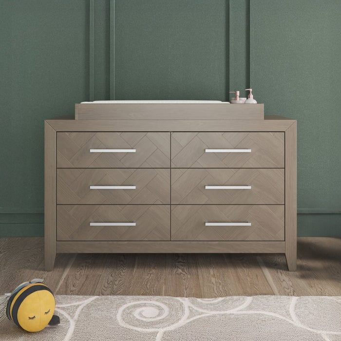 A six-drawer wooden dresser with silver handles stands against a green wall. On top sits the Child Craft F01792 Universal Dressing Kit in Crescent Grey, along with baby items; a plush bee toy rests on the patterned rug in front.