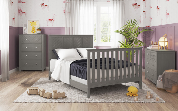 A cozy kids bedroom features the Child Craft F06401 Full-Size Bed Rail in Lunar Gray, with matching dresser and chest. Wooden toys and stuffed animals rest on a light rug. Soft pink and white walls, plants, and sunlit sheer curtains complete the scene.