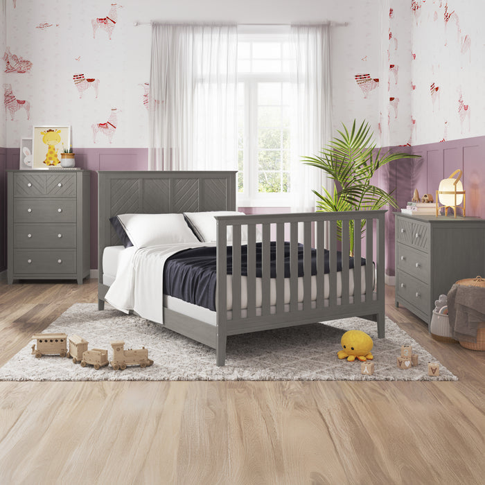 A modern kids’ room features gray Child Craft furniture, including a crib with the F06401 Full-Size Bed Rail in Lunar Gray, two dressers, and a bed. The space has wood floors, pink/white walls, stuffed toys, and a large window with white curtains.