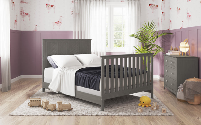 A cozy modern bedroom features the Child Craft F06401 Full-Size Bed Rail in Lunar Gray, white bedding, black blanket, light gray rug with wooden toys, purple walls, window, dresser, plant, playful wallpaper, and a handy crib conversion kit.
