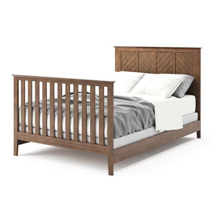 The Child Craft F06401 full-size bed rails in Cocoa Bean are shown with a wooden frame, chevron-patterned headboard, slatted footboard, and gray and white bedding with two pillows, viewed from a side angle.