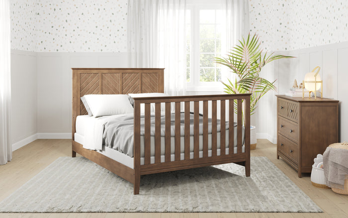 A bright bedroom features a wooden bed with Child Craft F06401 Full-Size Bed Rails in Cocoa Bean, white bedding, a matching dresser, a large potted plant, gray rug, white wainscoting, patterned wallpaper, and sunlight through sheer curtains.