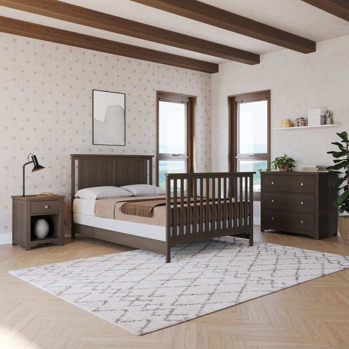 A modern bedroom features Child Craft’s F06401 Full-Size Bed Rails in Dapper Gray, a dark wood bed frame, nightstand, dresser, geometric rug, desk lamp, plant, light dot-patterned walls, and three windows for natural light.