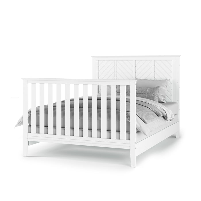 The Child Craft F06401 features full-size bed rails in matte white, a white wooden frame with slatted headboard and footboard, and is styled with gray and white bedding, pillows, and a neatly folded blanket.