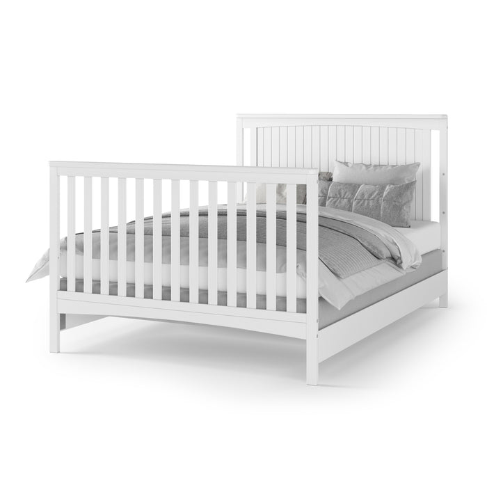 The Child Craft F06401 Full-Size Bed Rails in matte white, crafted from sturdy pine wood, add lasting durability to a white bed frame with vertical slats on the headboard and footboard, finished with gray and white bedding and pillows.