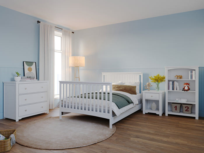 A bright bedroom with light blue walls, white furniture including the Child Craft F06401 Full-Size Bed Rails in matte white, a dresser, nightstand, bookshelf, standing lamp, round beige rug on wood floors, and a window with sheer curtains for natural light.