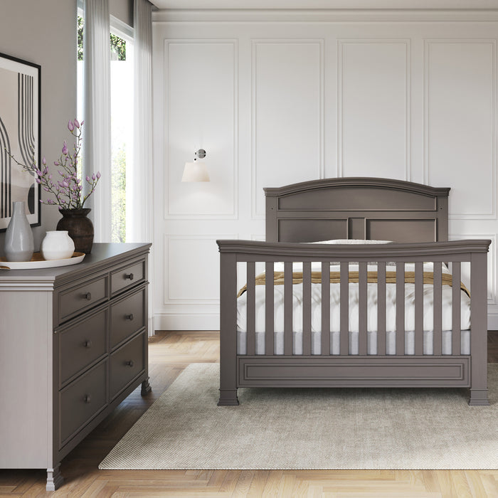 A nursery room features a Child Craft Chelsea Gray crib with F06424 full-size bed rails, plus a matching dresser. A beige rug sits on a light wood floor, framed art and vases decorate the dresser, and white paneled walls surround a large window.