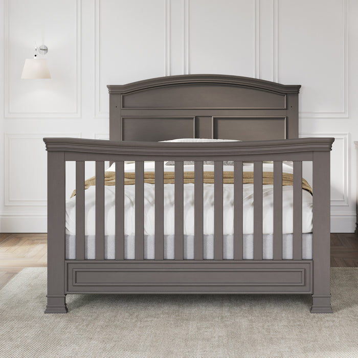 A gray wooden Child Craft crib with slatted sides is on a light rug in a room with white paneled walls, hardwood floors, and a wall lamp. Compatible with Child Craft F06424 Chelsea Gray Full-Size Bed Rails for future conversion.