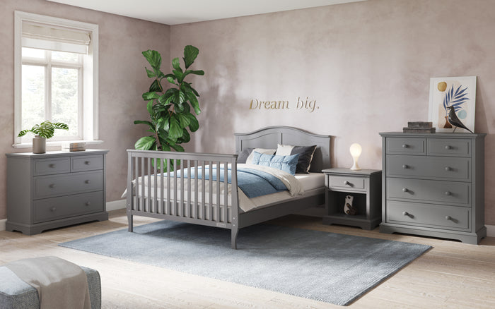 A cozy bedroom features gray furniture, including the Child Craft F06454 Full-Size Bed Rails in Cool Gray, a dresser, nightstand, and chest of drawers. A plant, lamp, and wall art decorate the room with "Dream big." above the bed.