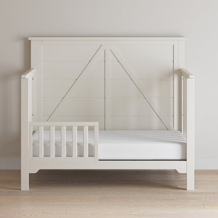 The Child Craft F09501 Toddler Guard Rail in Brushed Cotton is a white wooden rail with a paneled design, perfect for smooth crib-to-toddler bed transitions and added safety, shown against a light wall on a wooden floor.