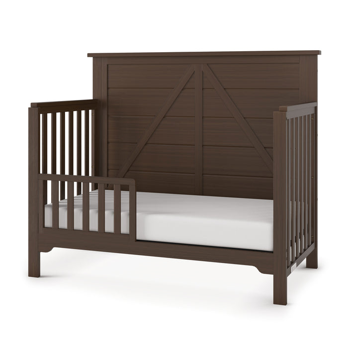 The Child Craft F09501 Toddler Guard Rail in Brushed Truffle is shown with slatted sides and a paneled backrest, paired with a white mattress—ideal for converting your crib into a toddler bed.