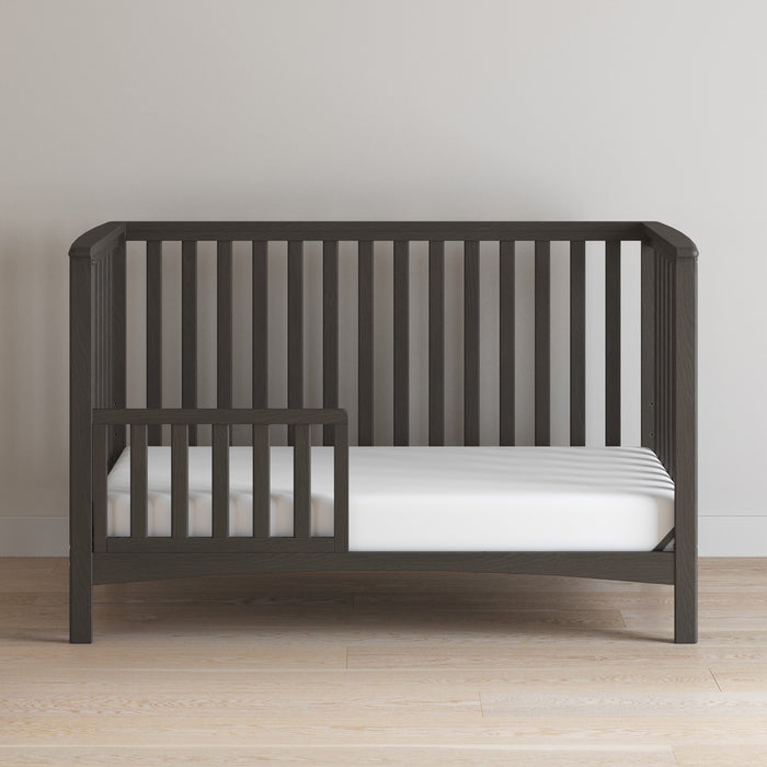 The Child Craft F09501 Toddler Guard Rail in Dapper Gray features sturdy wood construction and slatted sides, shown here with a white mattress (no bedding) on a light wood floor against a plain beige wall.