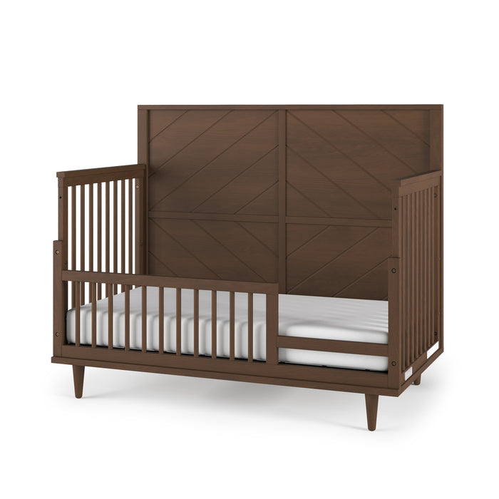 The Child Craft F09502 Toddler Guard Rail in Toasted Chestnut is attached to a wooden toddler bed with a high, paneled headboard, open slatted sides, and a white mattress, shown against a plain white background.