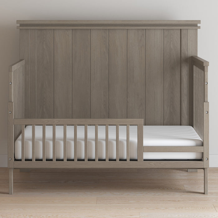 A Child Craft toddler bed with high paneled back and low side rails features the F09503 Crescent Grey Toddler Guard Rail, includes a white mattress, and is set on a light wood floor against a plain wall.